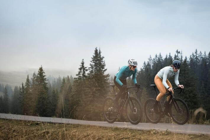 Ženska in moški kolesarita v termo kolesarskih oblačilih, v ozadju pokrajina in cesta.